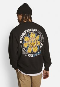Schwarzer Sweatshirt mit einem großen gelben Blumenmotiv auf der Rückseite, umgeben von dem Text "REDEFINED REBEL STREETWEAR."