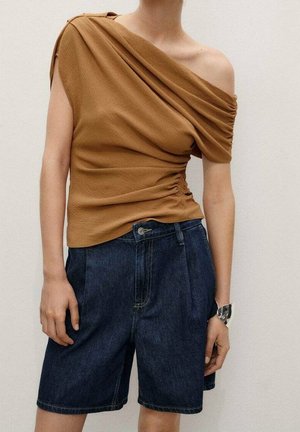 Person iført en rynket brun ene-skulder-top og mørke denim knælange shorts med et sølvarmbånd på håndleddet.