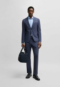 Completo blu navy con vestibilità slim, tessuto strutturato, abbinato a una camicia azzurra chiaro. Tiene una borsa nera strutturata e indossa scarpe nere lucide.