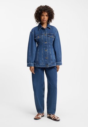 Kvinde med krøllet hår har en tætsiddende blå denimjakke med knapper, matchende løse denimbukser og brune og hvide mønstrede sko.