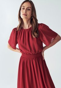 Crvena haljina s nabranim strukom, kratkim napuhanim rukavima i okruglim izrezom. Tkanina se čini laganom i lagano prozirnom, pada do sredine lista.