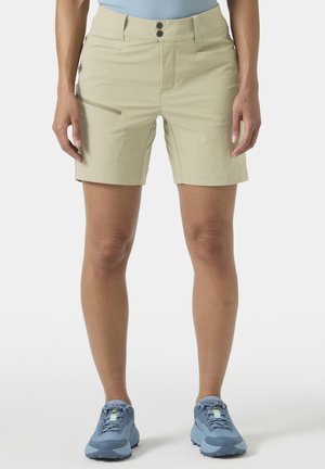 Mujer con pantalones cortos beige hasta la rodilla, con bolsillo lateral con cremallera, y zapatos deportivos azules, de pie frente a un fondo liso.