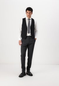 Schwarze Weste über einem weißen Hemd, kombiniert mit grauen Hosen und schwarzen Schuhen. Slim Fit-Design mit glatter Textur und fünfknopfiger Verschluss.