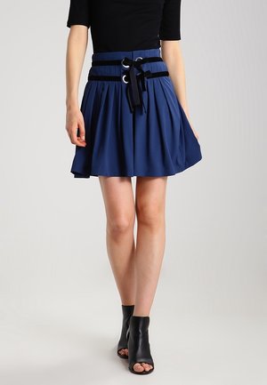 Femme portant une jupe bleue plissée avec des ceintures ruban noires et des bottines noires à bouts ouverts, debout devant un fond uni.