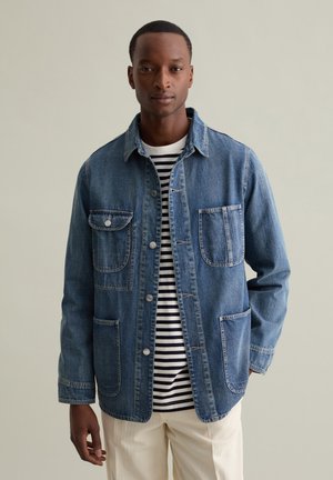 Denim jacket in medium blue with a collar, front button closure, two chest pockets, and long sleeves over a striped shirt. Smooth texture.