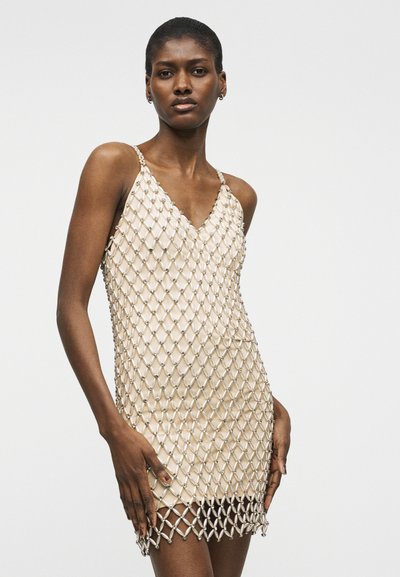 Γυναίκα που φοράει μπεζ φόρεμα-σλιπ με επικάλυψη από ασημένιο δίχτυ με χάντρες, όρθια μπροστά σε απλό φωτεινό φόντο.