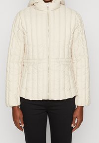 Pikowana kremowa kurtka puchowa z kapturem, z przodu zapinana na zamek błyskawiczny, z elastycznymi mankietami. Ma dopasowaną talię i gładką teksturę.