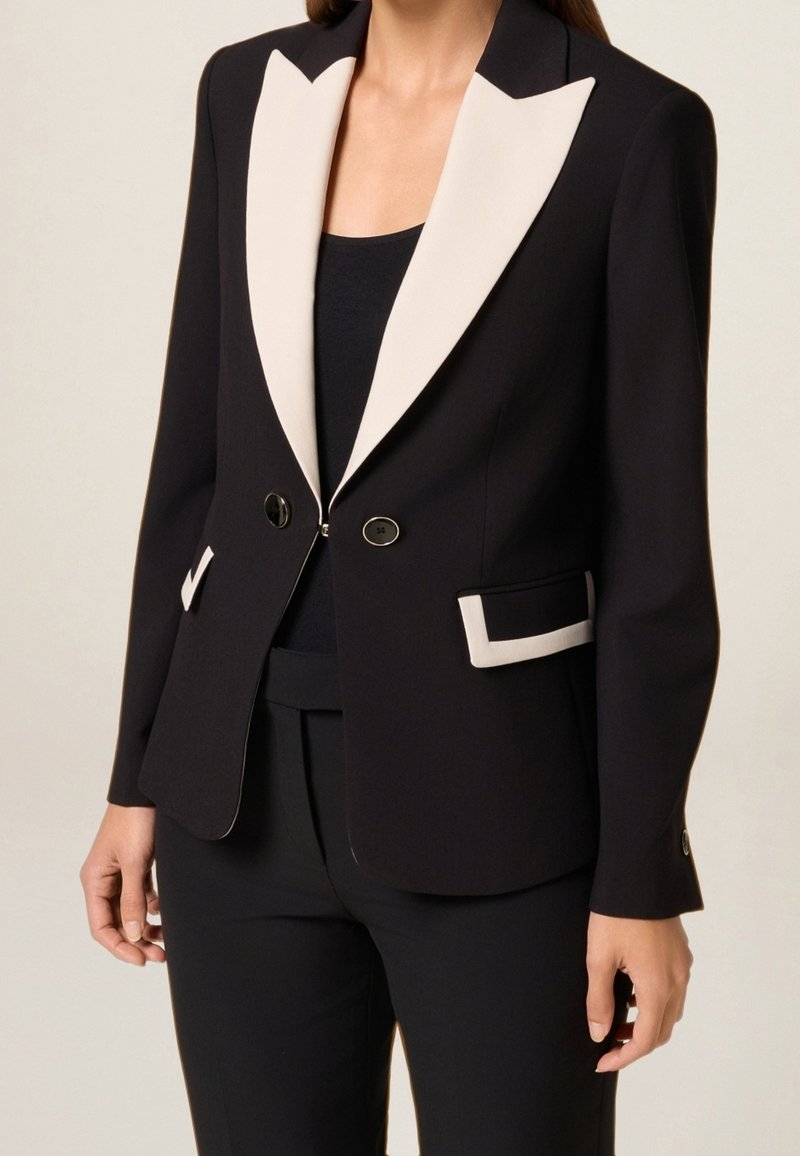 Femme portant un blazer noir ajusté avec des revers blancs contrastants et des bordures de poches, associé à un pantalon noir et un haut noir.