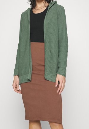 Femme portant un sweat à capuche zippé en maille verte, un top noir, une jupe crayon côtelée marron et un collier chaîne doré, debout devant un fond uni.
