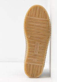 Semelle d'une chaussure en caoutchouc beige avec des motifs de grip texturés et le logo "Gabor", posée verticalement contre une surface blanche.