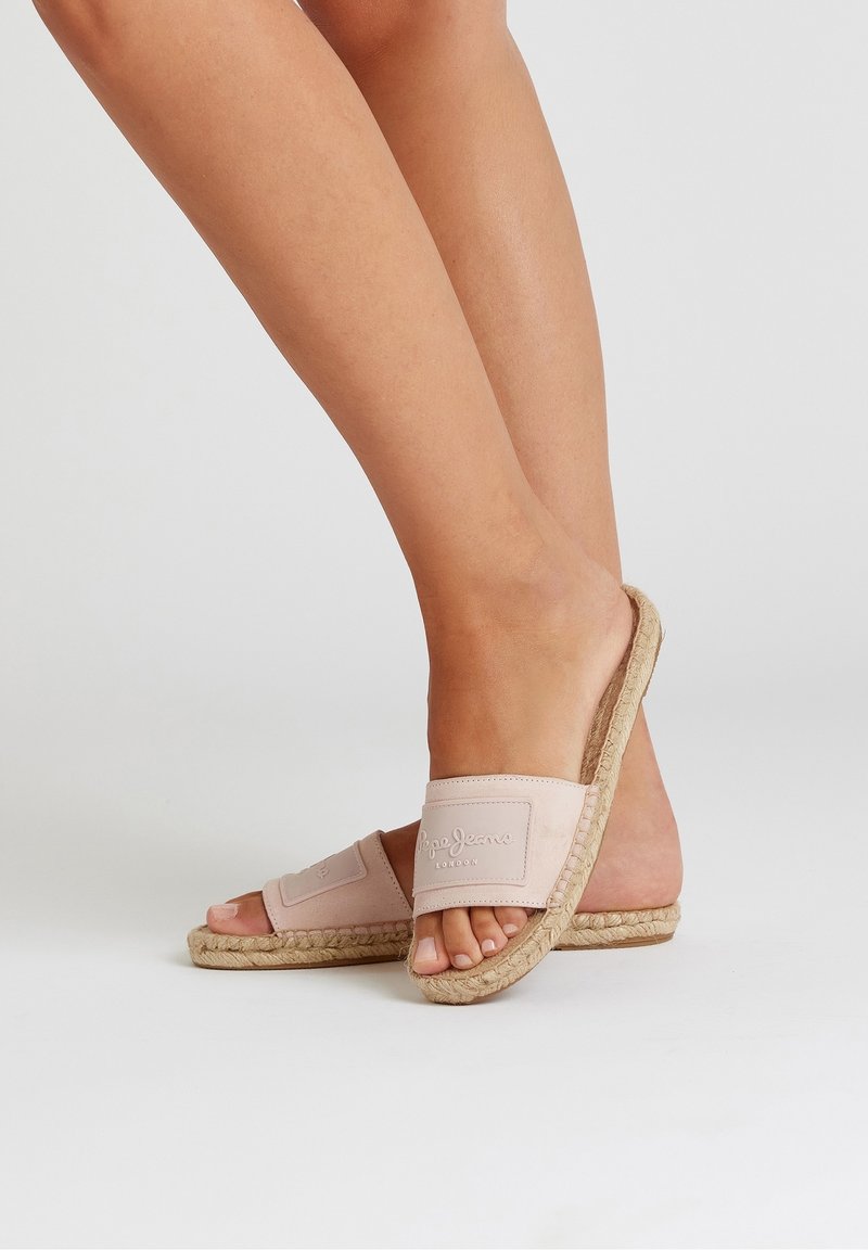Pieds nus portant des sandales espadrilles rose clair à semelles en corde tressée sur un fond clair uni.