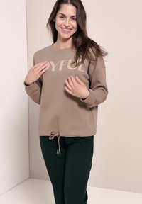 Kvinde med langt brunt hår, smilende, iført en beige sweatshirt med teksten "JOYFUL" og sorte bukser, står foran en ensfarvet baggrund.