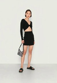 Top cropped preto de manga longa com frente torcida, saia mini preta de cintura alta, sandálias deslisantes pretas e uma pequena bolsa de ombro com padrão.