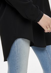 Czarna bluzka z długim rękawem o zaokrąglonym dolnym krawędzi, połączona z jasnoniebieskimi dżinsami. Materiał ma gładką teksturę i luźny krój rękawów.