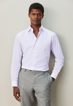 Bărbat cu păr scurt și creț, purtând o cămașă mov deschis cu mâneci lungi și pantaloni gri de costum, cu o mână în buzunar, pe un fundal simplu.