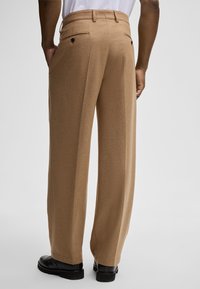 Spodnie o szerokich nogawkach w jasnobrązowym materiale, z tylnymi kieszeniami z czarnymi guzikami oraz eleganckim, dopasowanym wykończeniem.