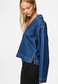 Zila džinsa pārmēness jaka ar V-veida kakla izgriezumu, garām piedurknēm, sānu šķirbām un kontrastējošām šuvēm. Strukturēts, neformāls dizains.