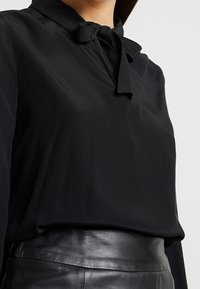Zwarte blouse van een gladde stof met een strikje bij de hals, lange mouwen en gecombineerd met een nauwsluitende zwarte leren rok.