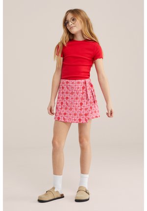 Fată cu păr lung și ochelari, purtând un tricou roșu cu mâneci scurte, fustă roz cu model, șosete albe și pantofi alunecători bej, stând calmă.