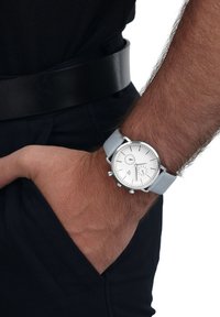Silberne Uhr mit weißem Ziffernblatt und hellgrauem Lederarmband, ausgestattet mit dünnen Stundenmarkierungen und Unterzifferblättern, am Handgelenk getragen gegen dunkle Kleidung.