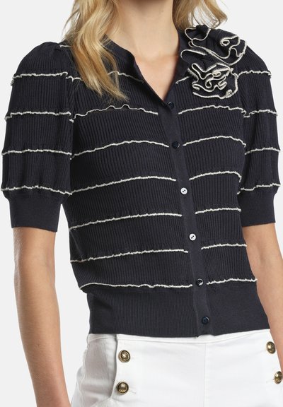 Cardigan scurt cu mâneci scurte, negru cu dungi orizontale albe, detaliu floral cu volănașe lângă umăr, asortat cu pantaloni albi cu nasturi aurii.