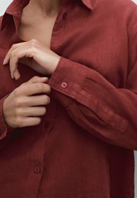 Koszulka z lnianego materiału w kolorze rdzy, z zapięciem na guziki przy kołnierzy, długimi rękawami z guziczkami oraz tkaniną o fakturze. Widać ręce regulujące mankiet.
