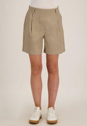 Beige, op maat gemaakte shorts met hoge taille en plooien, gedragen met witte sneakers bij een staande persoon tegen een neutrale achtergrond.