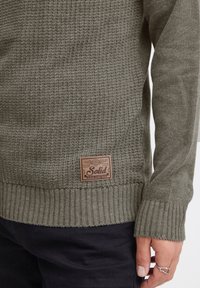 Olivgrüner, gestrickter Pullover mit gerippten Bündchen, versehen mit einem rechteckigen, braunen Lederpatch an der unteren Seite, das den embossed Markennamen und Details trägt.