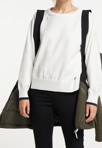 Pull en maille blanche avec col rond, ourlet côtelé et accents bleu marine aux poignets, associé à une doudoune vert foncé.