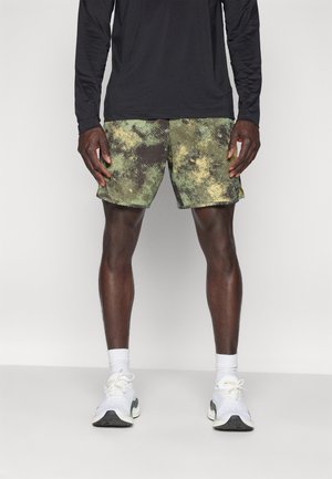 Man draagt groene en zwarte patroon kortebroek, zwarte top met lange mouwen, witte sokken en witte sportschoenen, staand op een lichte achtergrond.