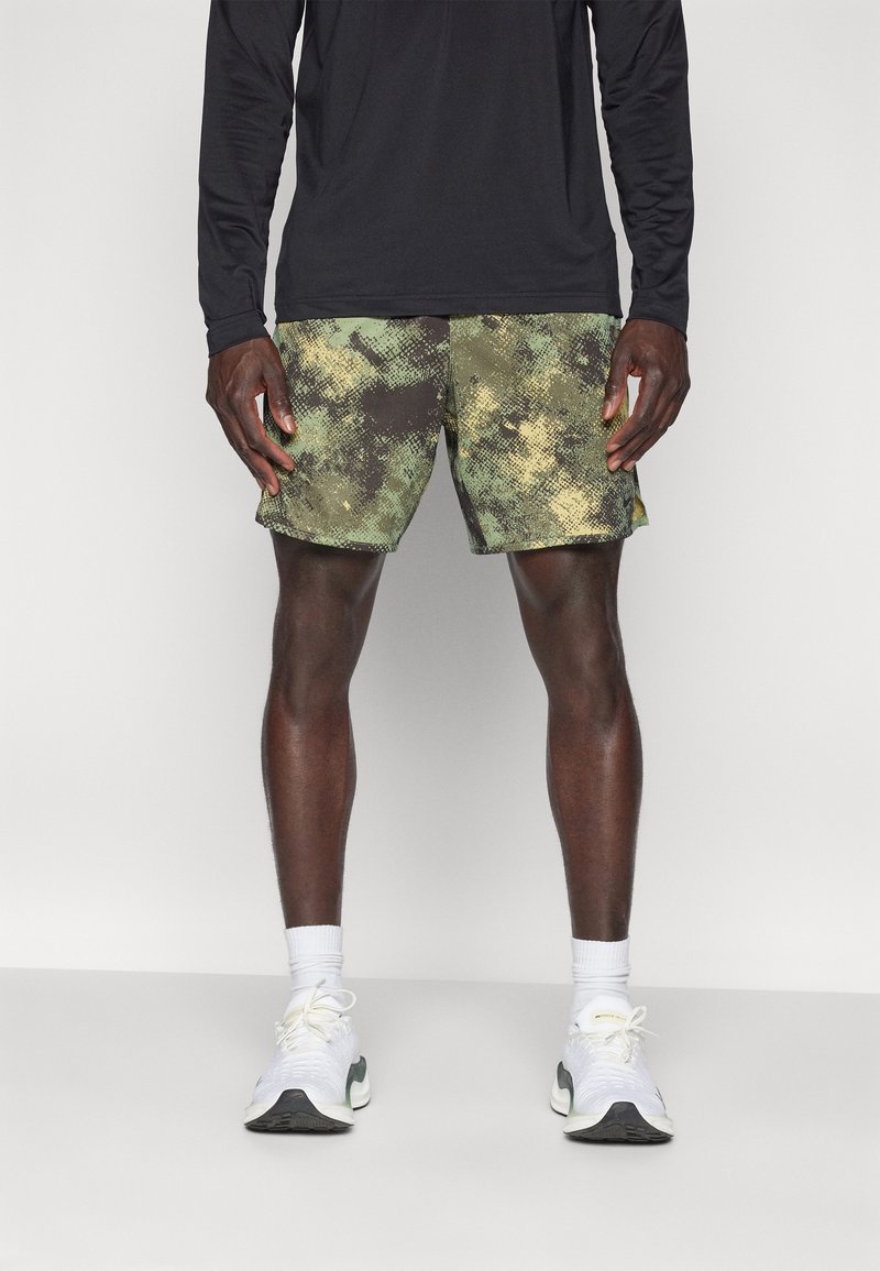Homme portant un short vert et noir à motifs, un haut noir à manches longues, des chaussettes blanches et des chaussures de sport blanches, debout sur un fond clair.