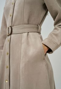 Abrigo de ante beige con cinturón, botones dorados y bolsillos laterales. Presenta un diseño ajustado y una textura suave.