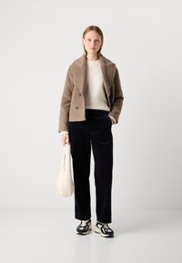 Vrouw staat in een brown cropped coat, witte sweater, zwarte wijde broek, zwart-witte sneakers, en houdt een witte gewatteerde handtas vast.