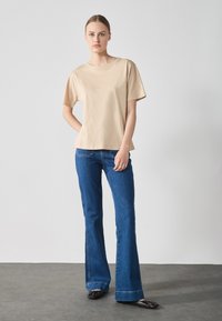 Beige Kurzarm-T-Shirt aus weichem Stoff, kombiniert mit blauen Schlagjeans im klassischen Fünf-Taschen-Design und schwarzen flachen Schuhen.