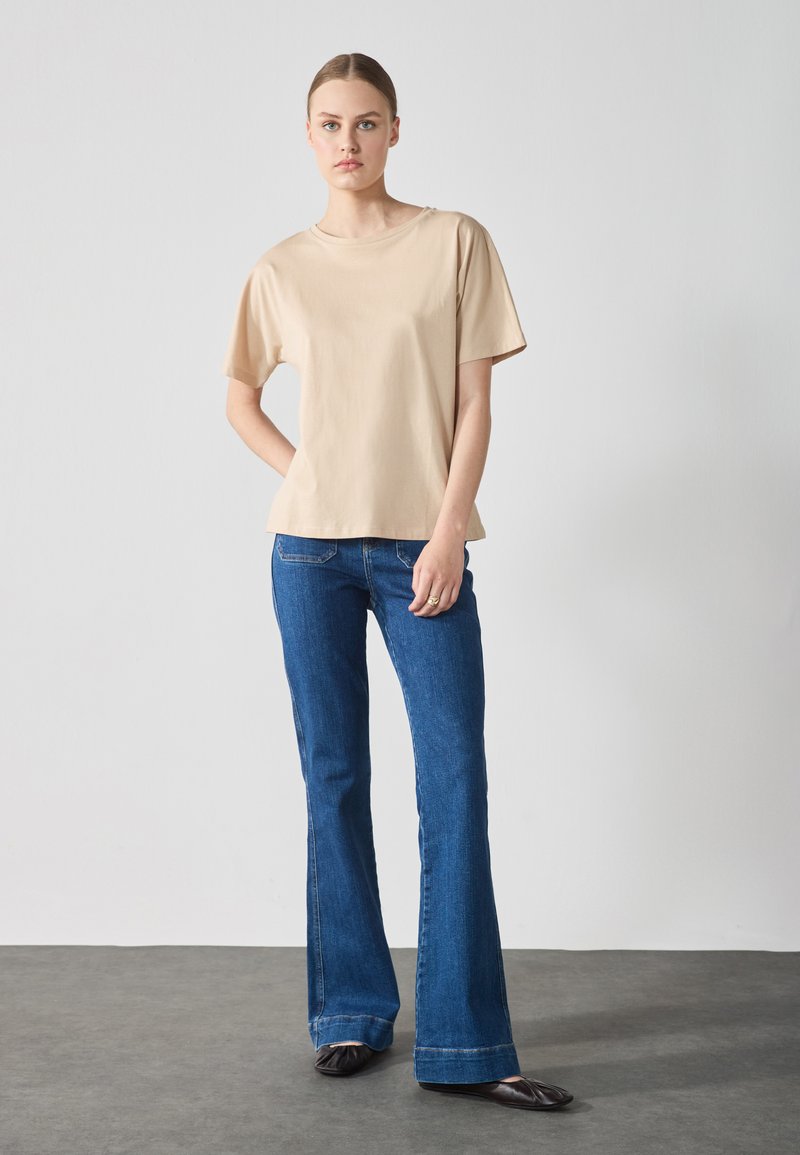 Beige Kurzarm-T-Shirt aus weichem Stoff, kombiniert mit blauen Schlagjeans im klassischen Fünf-Taschen-Design und schwarzen flachen Schuhen.
