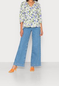 Blouse à imprimé floral avec manches 3/4, fond blanc, fleurs bleues et vertes ; jean en denim bleu taille haute à jambes larges ; sandales à talons orange.
