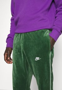 Nike fløjlsbukser i mørkegrøn med hvide side striber, med et broderet logo. Kombineret med en ensfarvet lilla sweatshirt.