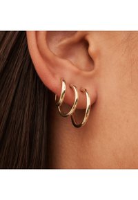 Boucles d'oreilles en or, trois empilées de tailles différentes, texture lisse, finition réfléchissante, accrochées à un lobe percé, sur fond de cheveux bruns.