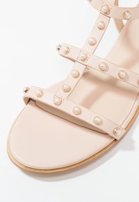 Sandale beige avec plusieurs brides ornées de clous ronds nacrés, semelle plate et design minimaliste.