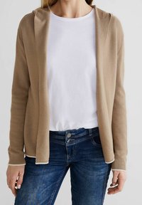 Beige stickad kofta med öppen front och huva, matchad med en vit t-shirt och blå jeans. Koftan har ribbade manschetter och nederkant.