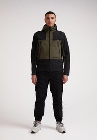Giacca impermeabile in verde oliva e nero con cappuccio, tasche con zip e design a pannelli a contrasto, abbinata a pantaloni cargo neri e scarpe.