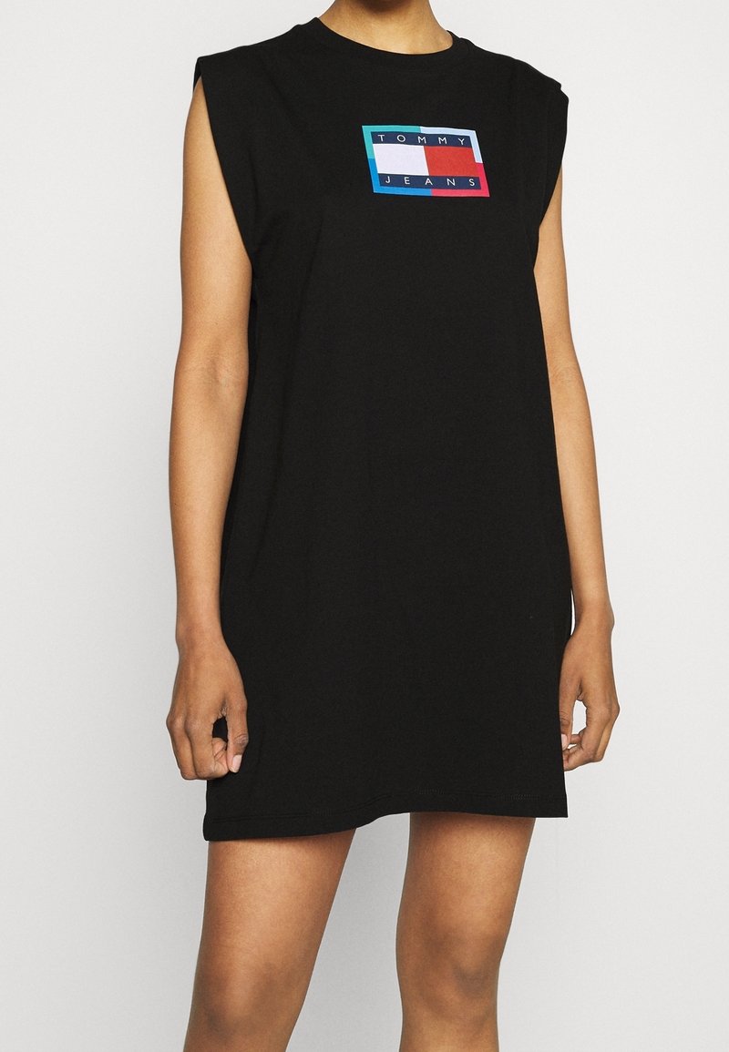 Czarna sukienka bez rękawów wykonana z bawełny, z prostokątną grafiką z napisem "TOMMY JEANS" w kolorach niebieskim, czerwonym i białym.