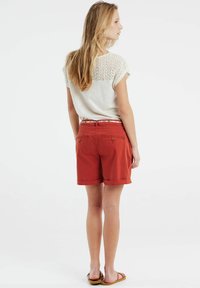Witte top met kanten detail op de rug, rode shorts met omgeslagen zoom en twee achterzakken, en bruine sandalen.