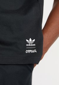 Camiseta de algodón negra con el logotipo blanco de Adidas Trefoil y el texto "PABLO VITTAR". Textura suave con cuello redondo y mangas cortas.
