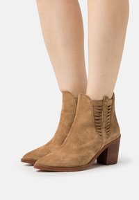 Kankaalikorkuiset vaaleanruskeat mokka-saappaat, joissa on paksut korkokengät ja kudotut sivupaneelit, käytettyinä paljaille jaloille valkoista taustaa vasten.