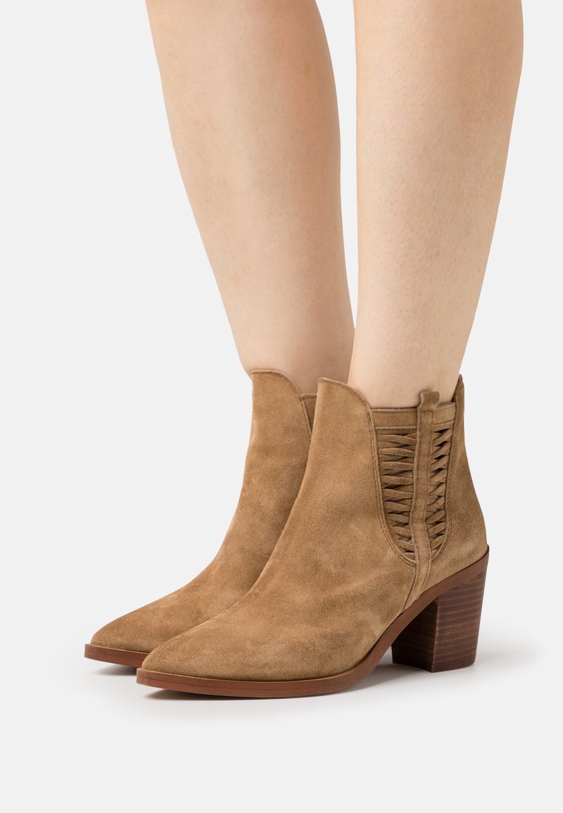 Kankaalikorkuiset vaaleanruskeat mokka-saappaat, joissa on paksut korkokengät ja kudotut sivupaneelit, käytettyinä paljaille jaloille valkoista taustaa vasten.