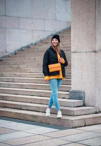 Černá zateplená bunda, žlutý svetr, světle modré džíny a bílé tenisky. Drží oranžovou kabelku. Stojí na kamenných schodech.