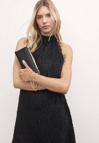 Juoda suknelė aukštu kaklu su tekstūruotais fringe. Modelis laiko stačiakampį juodą rankinuką su auksinėmis detalėmis ir dėvi sluoksniuotas auksines apyrankes.