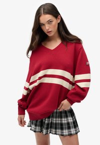 Rød strikket sweater med hvide striber, v-hals og ribkant, parret med en sort-hvid ternet plisseret nederdel.