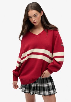 Czerwony sweter z dzianiny z białymi paskami, w serek, z ribbowanymi mankietami, zestawiony z plisowaną spódnicą w czarno-białą kratę.